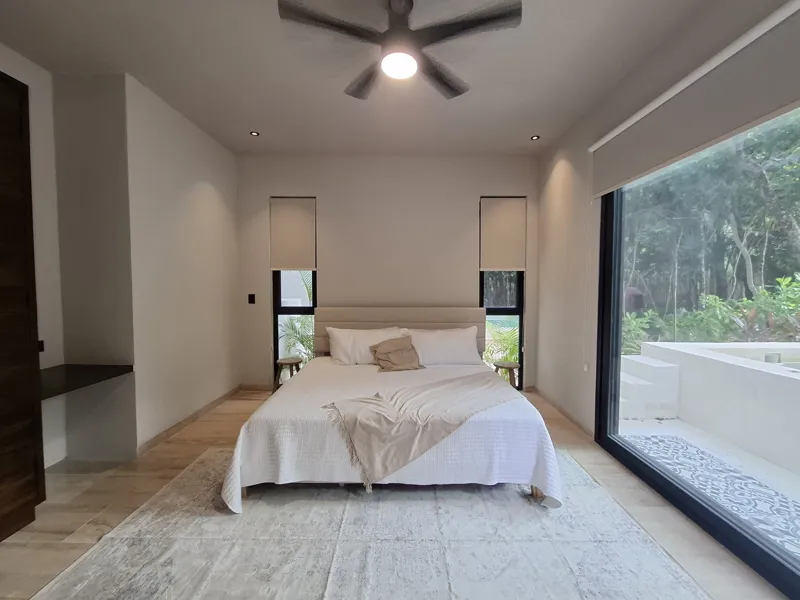 A modern kitchen and dining area in an Attha Holistika 1-bedroom unit, featuring light wood cabinetry, a stainless steel refrigerator, a black countertop, a round wooden dining table with four chairs, and a ceiling fan, with a large window offerin...