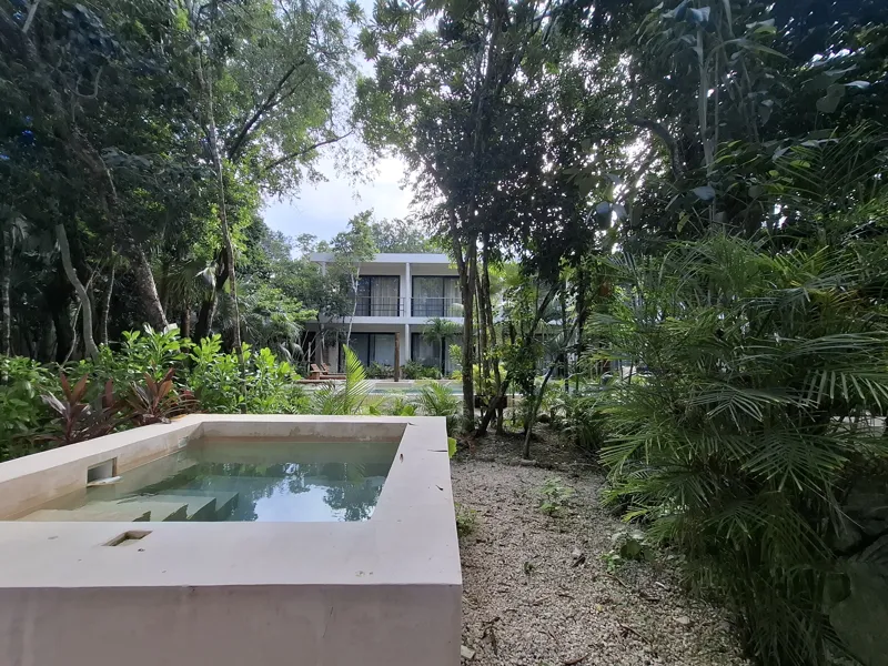 A modern kitchen and dining area in an Attha Holistika 1-bedroom unit, featuring light wood cabinetry, a stainless steel refrigerator, a black countertop, a round wooden dining table with four chairs, and a ceiling fan, with a large window offerin...