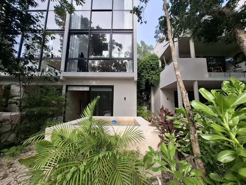 A modern kitchen and dining area in an Attha Holistika 1-bedroom unit, featuring light wood cabinetry, a stainless steel refrigerator, a black countertop, a round wooden dining table with four chairs, and a ceiling fan, with a large window offerin...