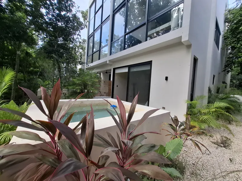 A modern kitchen and dining area in an Attha Holistika 1-bedroom unit, featuring light wood cabinetry, a stainless steel refrigerator, a black countertop, a round wooden dining table with four chairs, and a ceiling fan, with a large window offerin...