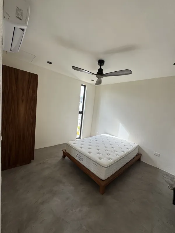 A wide shot of a finished modern apartment interior at Gold Garden, featuring a compact kitchen with concrete countertops, wooden cabinets, and a stainless steel refrigerator, opening into a living area with concrete floors, white walls, and large...