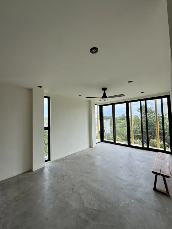 A wide shot of a finished modern apartment interior at Gold Garden, featuring a compact kitchen with concrete countertops, wooden cabinets, and a stainless steel refrigerator, opening into a living area with concrete floors, white walls, and large...