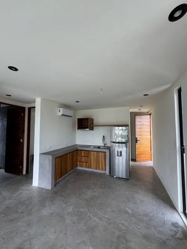 A wide shot of a finished modern apartment interior at Gold Garden, featuring a compact kitchen with concrete countertops, wooden cabinets, and a stainless steel refrigerator, opening into a living area with concrete floors, white walls, and large...
