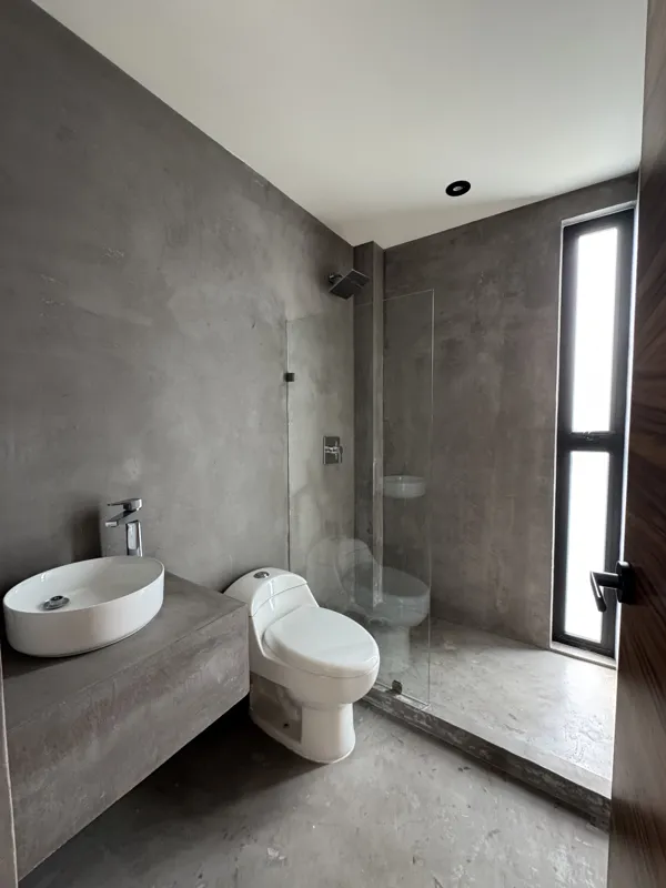 A wide shot of a finished modern apartment interior at Gold Garden, featuring a compact kitchen with concrete countertops, wooden cabinets, and a stainless steel refrigerator, opening into a living area with concrete floors, white walls, and large...