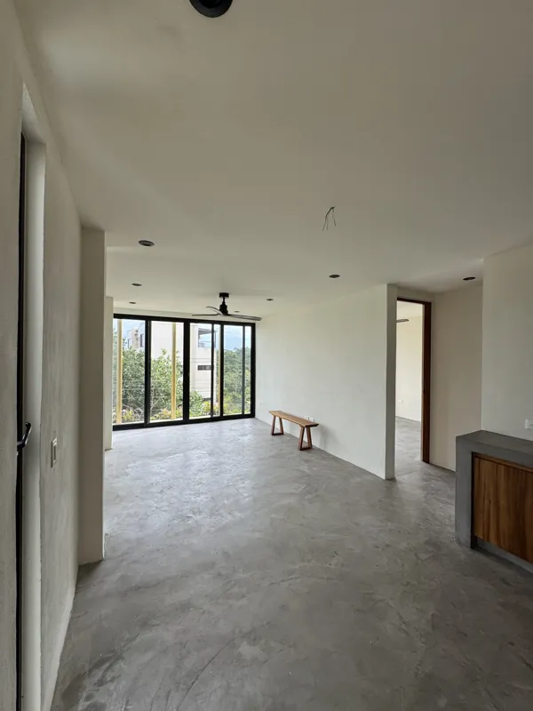 A wide shot of a finished modern apartment interior at Gold Garden, featuring a compact kitchen with concrete countertops, wooden cabinets, and a stainless steel refrigerator, opening into a living area with concrete floors, white walls, and large...
