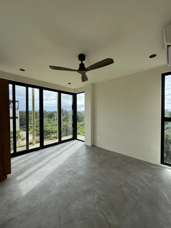 A wide shot of a finished modern apartment interior at Gold Garden, featuring a compact kitchen with concrete countertops, wooden cabinets, and a stainless steel refrigerator, opening into a living area with concrete floors, white walls, and large...