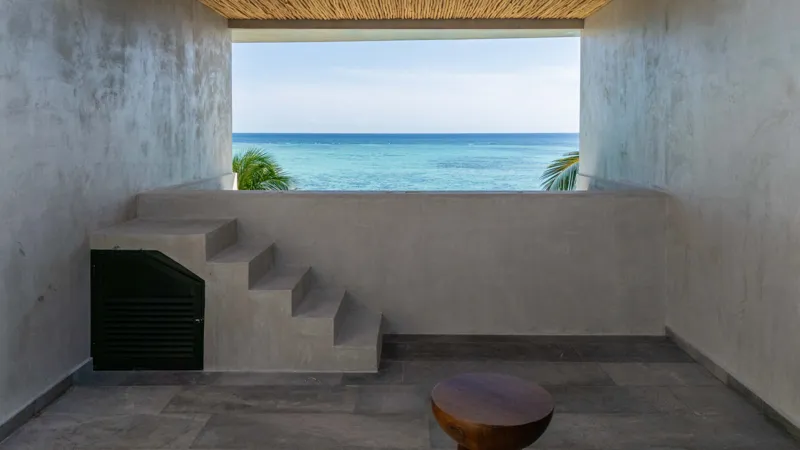 High-angle aerial shot of the NEEA development, a contemporary multi-story building with a rectangular rooftop pool, positioned directly on a white sand beach. The clear turquoise waters of Bahía Soliman stretch out, and numerous vibrant green pal...