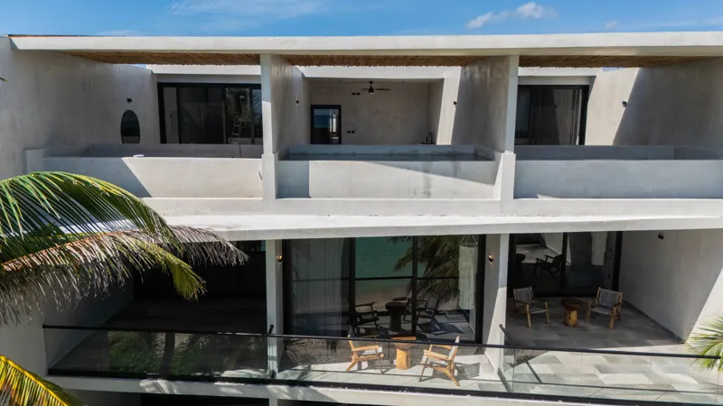 High-angle aerial shot of the NEEA development, a contemporary multi-story building with a rectangular rooftop pool, positioned directly on a white sand beach. The clear turquoise waters of Bahía Soliman stretch out, and numerous vibrant green pal...