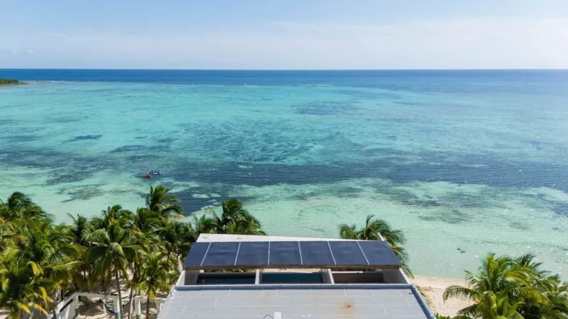 High-angle aerial shot of the NEEA development, a contemporary multi-story building with a rectangular rooftop pool, positioned directly on a white sand beach. The clear turquoise waters of Bahía Soliman stretch out, and numerous vibrant green pal...