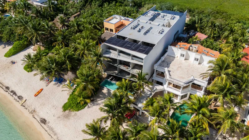 High-angle aerial shot of the NEEA development, a contemporary multi-story building with a rectangular rooftop pool, positioned directly on a white sand beach. The clear turquoise waters of Bahía Soliman stretch out, and numerous vibrant green pal...
