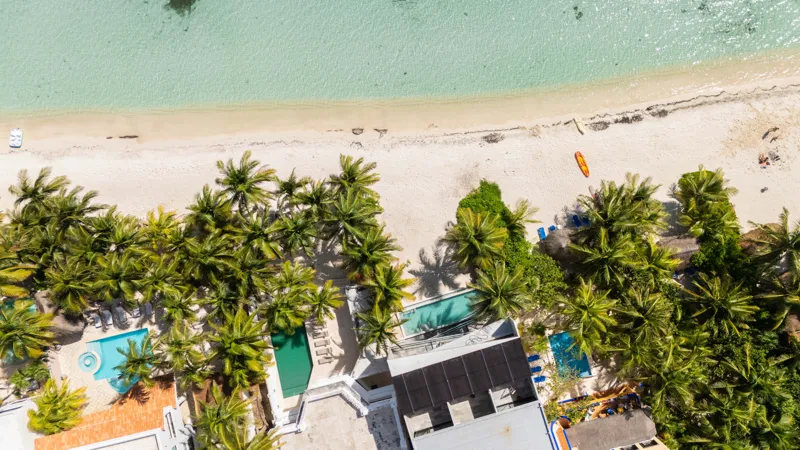 High-angle aerial shot of the NEEA development, a contemporary multi-story building with a rectangular rooftop pool, positioned directly on a white sand beach. The clear turquoise waters of Bahía Soliman stretch out, and numerous vibrant green pal...