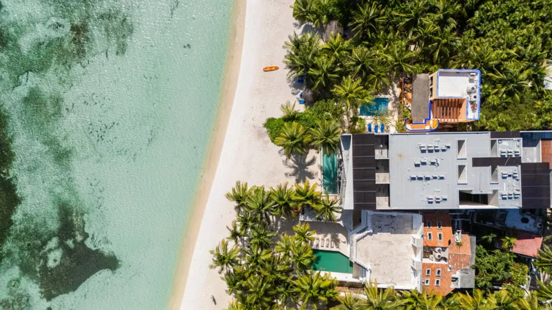 High-angle aerial shot of the NEEA development, a contemporary multi-story building with a rectangular rooftop pool, positioned directly on a white sand beach. The clear turquoise waters of Bahía Soliman stretch out, and numerous vibrant green pal...
