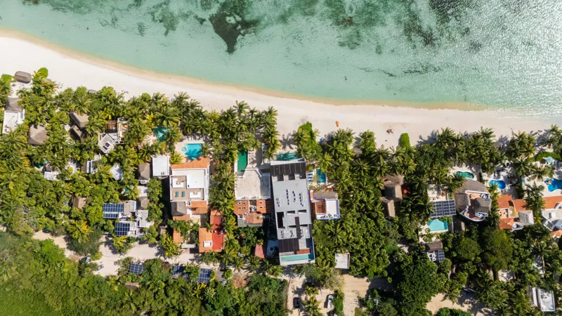 High-angle aerial shot of the NEEA development, a contemporary multi-story building with a rectangular rooftop pool, positioned directly on a white sand beach. The clear turquoise waters of Bahía Soliman stretch out, and numerous vibrant green pal...