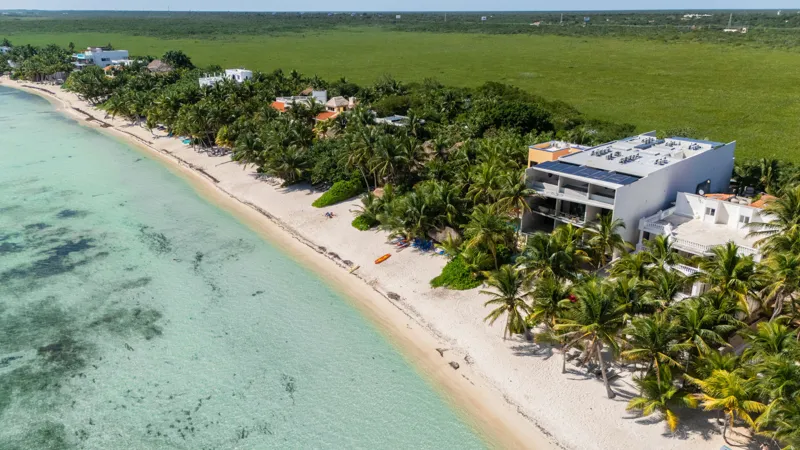 High-angle aerial shot of the NEEA development, a contemporary multi-story building with a rectangular rooftop pool, positioned directly on a white sand beach. The clear turquoise waters of Bahía Soliman stretch out, and numerous vibrant green pal...
