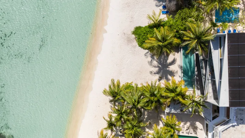 High-angle aerial shot of the NEEA development, a contemporary multi-story building with a rectangular rooftop pool, positioned directly on a white sand beach. The clear turquoise waters of Bahía Soliman stretch out, and numerous vibrant green pal...