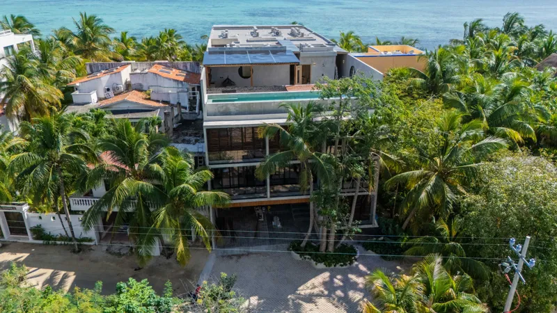 High-angle aerial shot of the NEEA development, a contemporary multi-story building with a rectangular rooftop pool, positioned directly on a white sand beach. The clear turquoise waters of Bahía Soliman stretch out, and numerous vibrant green pal...