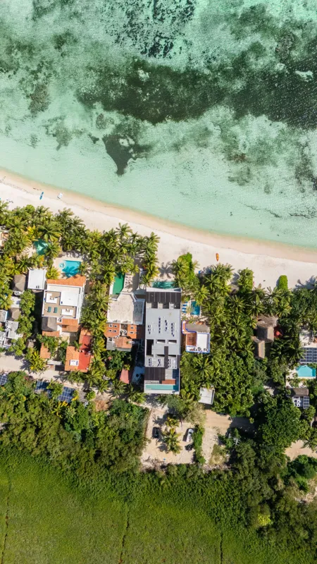 High-angle aerial shot of the NEEA development, a contemporary multi-story building with a rectangular rooftop pool, positioned directly on a white sand beach. The clear turquoise waters of Bahía Soliman stretch out, and numerous vibrant green pal...