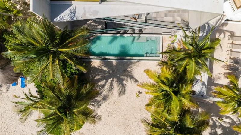 High-angle aerial shot of the NEEA development, a contemporary multi-story building with a rectangular rooftop pool, positioned directly on a white sand beach. The clear turquoise waters of Bahía Soliman stretch out, and numerous vibrant green pal...