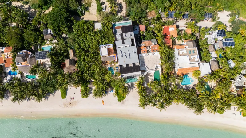 High-angle aerial shot of the NEEA development, a contemporary multi-story building with a rectangular rooftop pool, positioned directly on a white sand beach. The clear turquoise waters of Bahía Soliman stretch out, and numerous vibrant green pal...