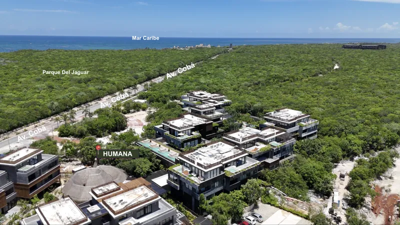 An aerial photograph of the Humana residential development in Tulum, Mexico, showing multiple multi-story buildings with flat roofs and integrated greenery, nestled within a dense jungle landscape. The Caribbean Sea is visible in the background, a...