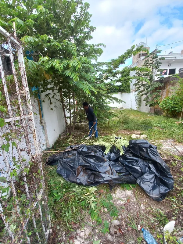 A man in a dark shirt and blue jeans crouches near an overgrown wall with utility meters, actively clearing weeds and debris from the Centurmex 25 development site, with a pickaxe resting on the ground nearby, indicating the start of site preparat...