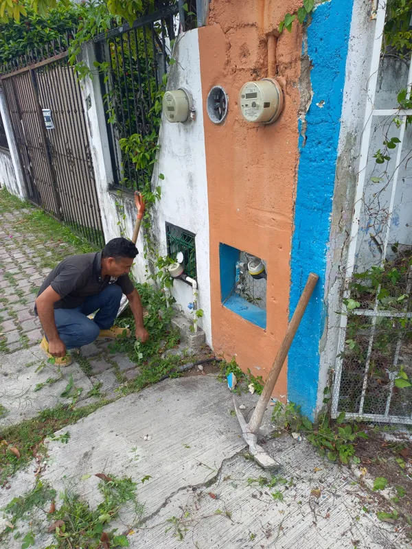 A man in a dark shirt and blue jeans crouches near an overgrown wall with utility meters, actively clearing weeds and debris from the Centurmex 25 development site, with a pickaxe resting on the ground nearby, indicating the start of site preparat...