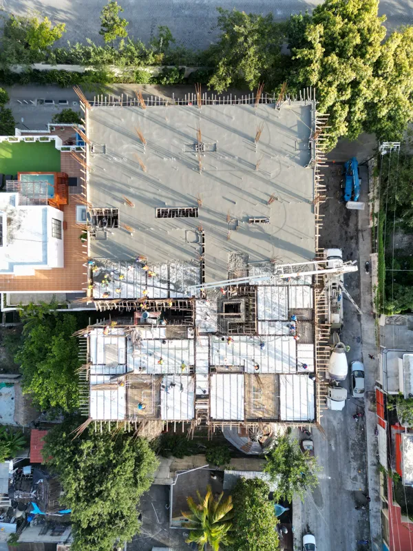 Aerial top-down view of the Maia development construction site, showing a large section of recently poured concrete on an upper floor, with rebar protruding for future levels. Below, workers in hard hats are active on lower levels where formwork a...