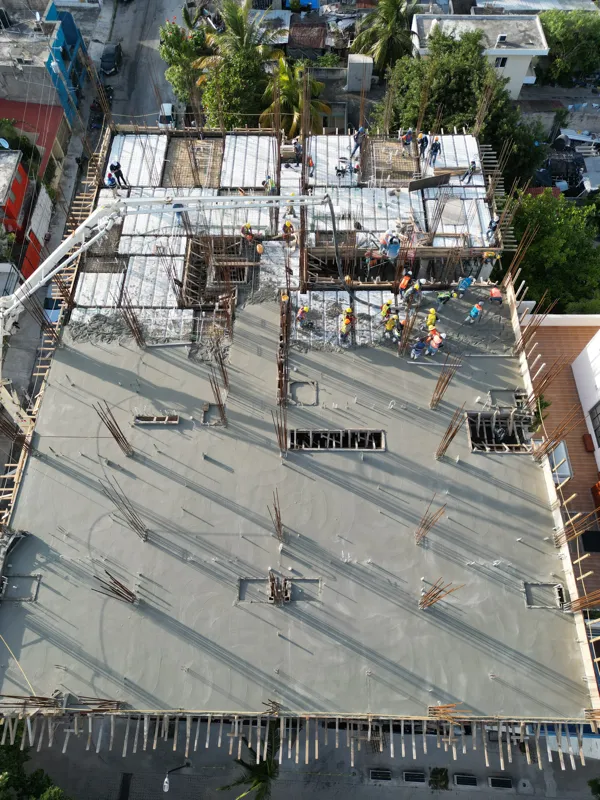Aerial top-down view of the Maia development construction site, showing a large section of recently poured concrete on an upper floor, with rebar protruding for future levels. Below, workers in hard hats are active on lower levels where formwork a...