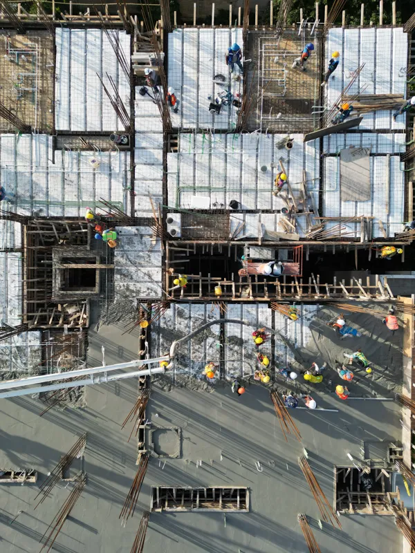 Aerial top-down view of the Maia development construction site, showing a large section of recently poured concrete on an upper floor, with rebar protruding for future levels. Below, workers in hard hats are active on lower levels where formwork a...