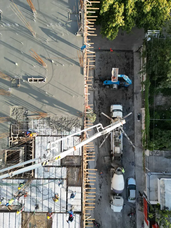 Aerial top-down view of the Maia development construction site, showing a large section of recently poured concrete on an upper floor, with rebar protruding for future levels. Below, workers in hard hats are active on lower levels where formwork a...
