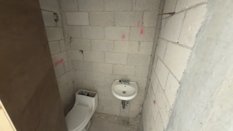 Interior view of a Studio34 apartment unit under construction, showing bare concrete block walls, an unfinished concrete floor, exposed electrical conduits on the ceiling, and a partially installed wooden door frame leading to another room. Red ma...