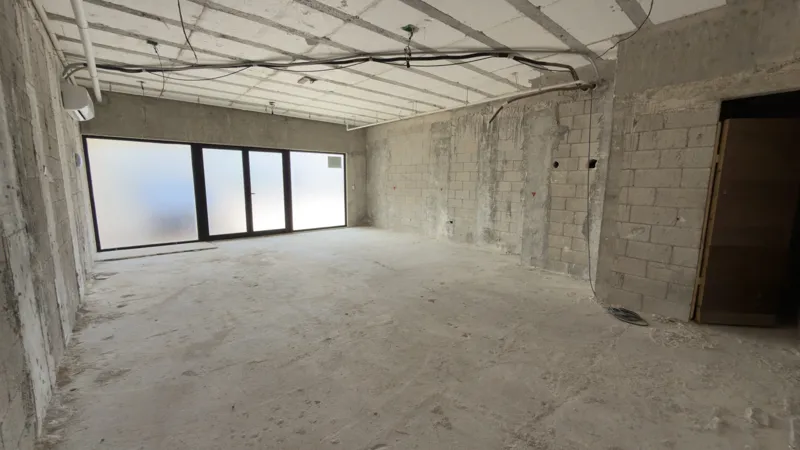 Interior view of a Studio34 apartment unit under construction, showing bare concrete block walls, an unfinished concrete floor, exposed electrical conduits on the ceiling, and a partially installed wooden door frame leading to another room. Red ma...