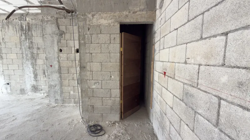 Interior view of a Studio34 apartment unit under construction, showing bare concrete block walls, an unfinished concrete floor, exposed electrical conduits on the ceiling, and a partially installed wooden door frame leading to another room. Red ma...