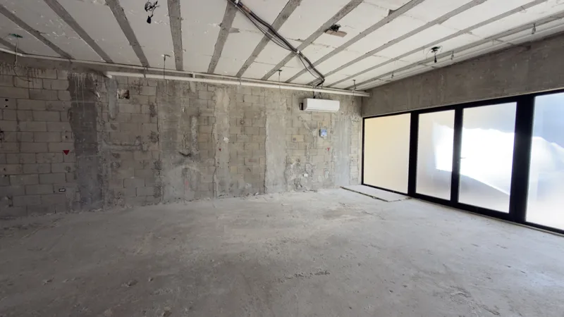 Interior view of a Studio34 apartment unit under construction, showing bare concrete block walls, an unfinished concrete floor, exposed electrical conduits on the ceiling, and a partially installed wooden door frame leading to another room. Red ma...