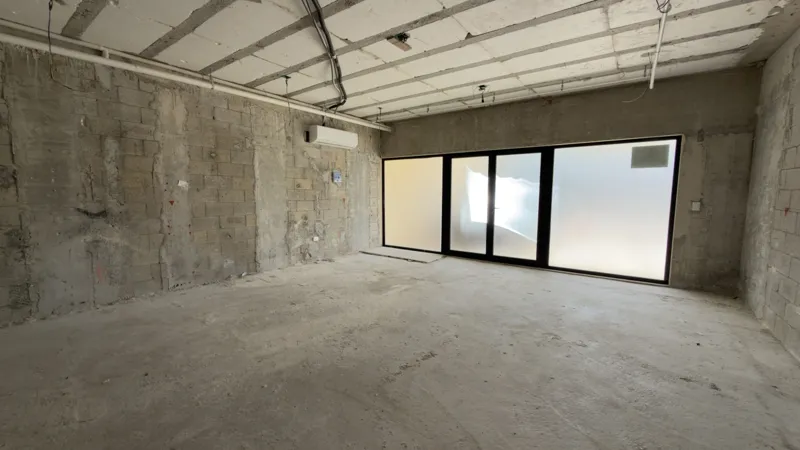 Interior view of a Studio34 apartment unit under construction, showing bare concrete block walls, an unfinished concrete floor, exposed electrical conduits on the ceiling, and a partially installed wooden door frame leading to another room. Red ma...