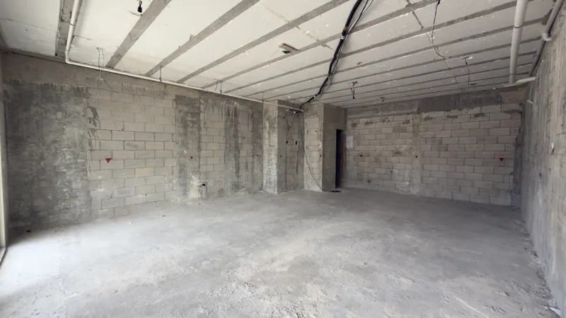 Interior view of a Studio34 apartment unit under construction, showing bare concrete block walls, an unfinished concrete floor, exposed electrical conduits on the ceiling, and a partially installed wooden door frame leading to another room. Red ma...