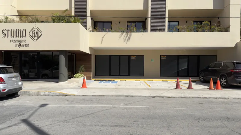 Interior view of a Studio34 apartment unit under construction, showing bare concrete block walls, an unfinished concrete floor, exposed electrical conduits on the ceiling, and a partially installed wooden door frame leading to another room. Red ma...