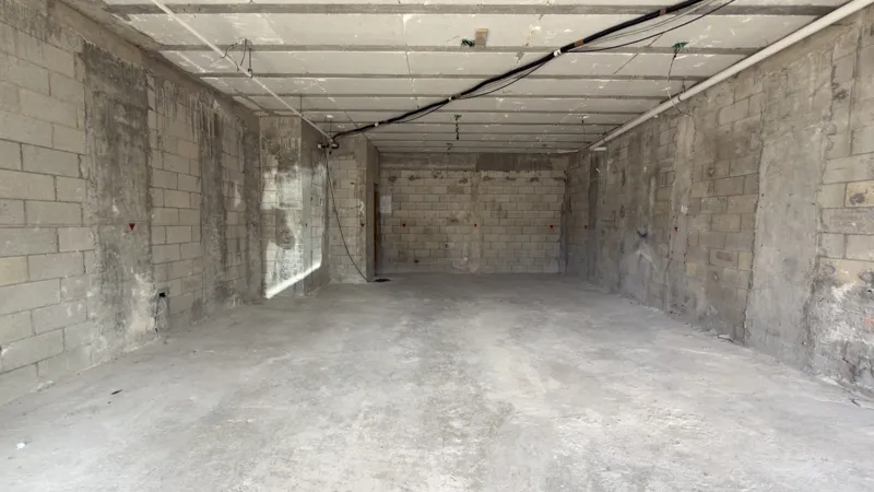 Interior view of a Studio34 apartment unit under construction, showing bare concrete block walls, an unfinished concrete floor, exposed electrical conduits on the ceiling, and a partially installed wooden door frame leading to another room. Red ma...