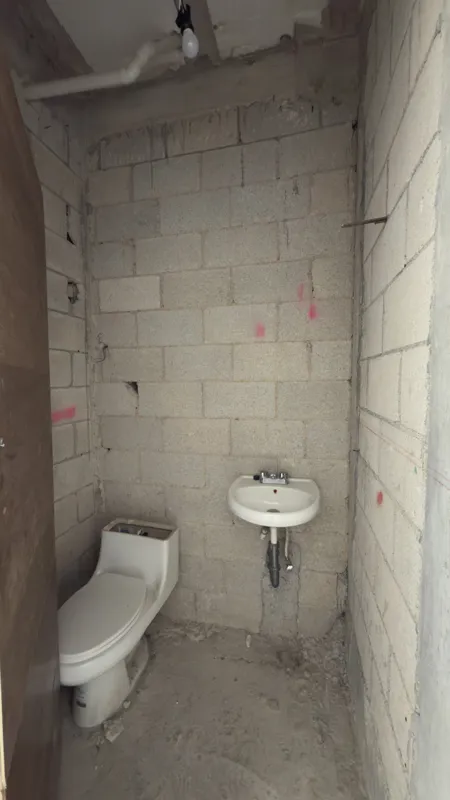 Interior view of a Studio34 apartment unit under construction, showing bare concrete block walls, an unfinished concrete floor, exposed electrical conduits on the ceiling, and a partially installed wooden door frame leading to another room. Red ma...