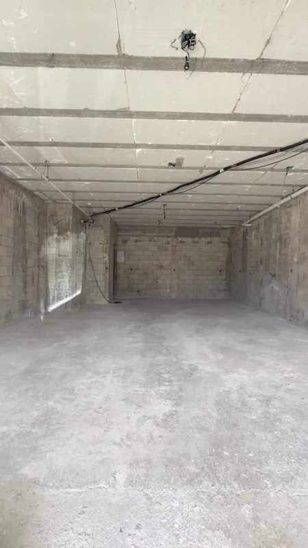 Interior view of a Studio34 apartment unit under construction, showing bare concrete block walls, an unfinished concrete floor, exposed electrical conduits on the ceiling, and a partially installed wooden door frame leading to another room. Red ma...