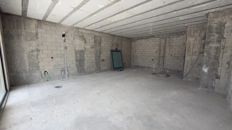 A full exterior view of the Studio34 development, a six-story modern building with a beige facade, dark vertical accents, and balconies, appearing largely complete under a clear blue sky. Orange traffic cones are visible on the street in front, an...