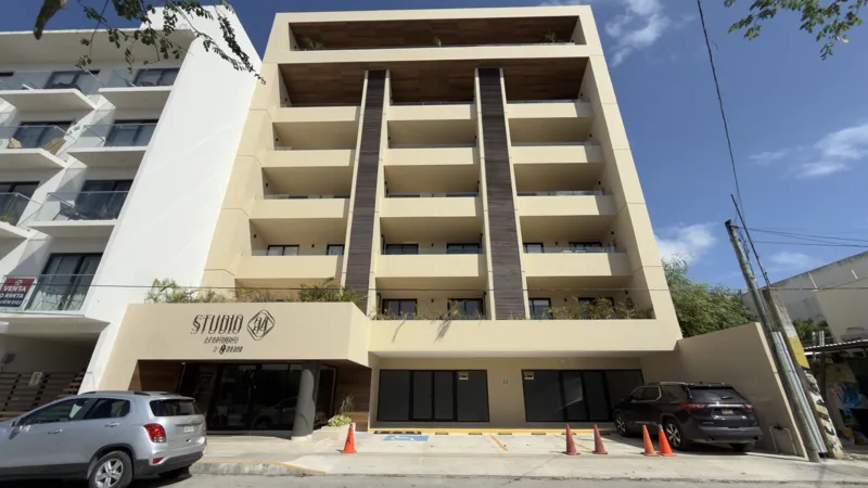 A full exterior view of the Studio34 development, a six-story modern building with a beige facade, dark vertical accents, and balconies, appearing largely complete under a clear blue sky. Orange traffic cones are visible on the street in front, an...