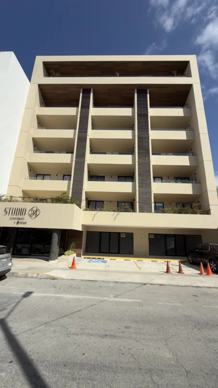 A full exterior view of the Studio34 development, a six-story modern building with a beige facade, dark vertical accents, and balconies, appearing largely complete under a clear blue sky. Orange traffic cones are visible on the street in front, an...
