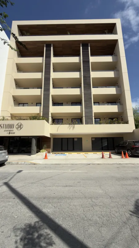 A full exterior view of the Studio34 development, a six-story modern building with a beige facade, dark vertical accents, and balconies, appearing largely complete under a clear blue sky. Orange traffic cones are visible on the street in front, an...