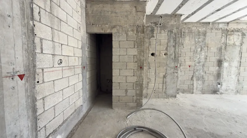 A full exterior view of the Studio34 development, a six-story modern building with a beige facade, dark vertical accents, and balconies, appearing largely complete under a clear blue sky. Orange traffic cones are visible on the street in front, an...