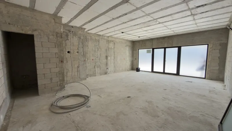 A full exterior view of the Studio34 development, a six-story modern building with a beige facade, dark vertical accents, and balconies, appearing largely complete under a clear blue sky. Orange traffic cones are visible on the street in front, an...