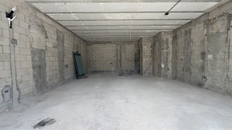 A full exterior view of the Studio34 development, a six-story modern building with a beige facade, dark vertical accents, and balconies, appearing largely complete under a clear blue sky. Orange traffic cones are visible on the street in front, an...