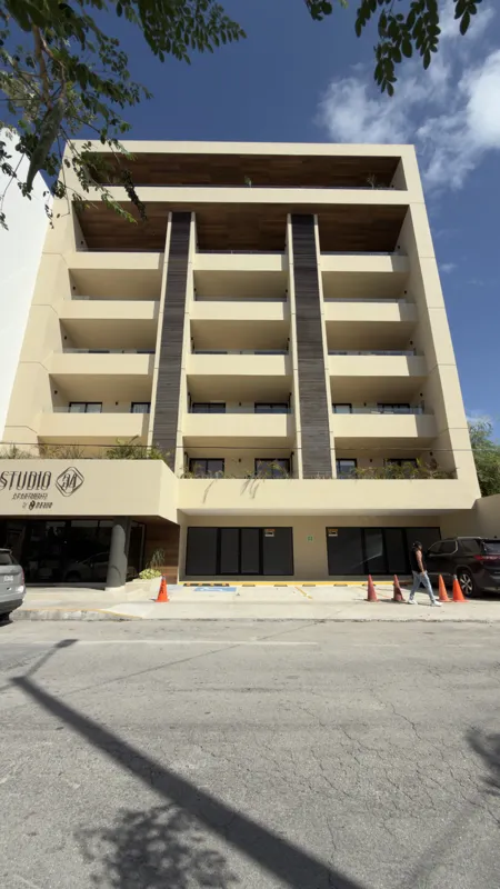 A full exterior view of the Studio34 development, a six-story modern building with a beige facade, dark vertical accents, and balconies, appearing largely complete under a clear blue sky. Orange traffic cones are visible on the street in front, an...