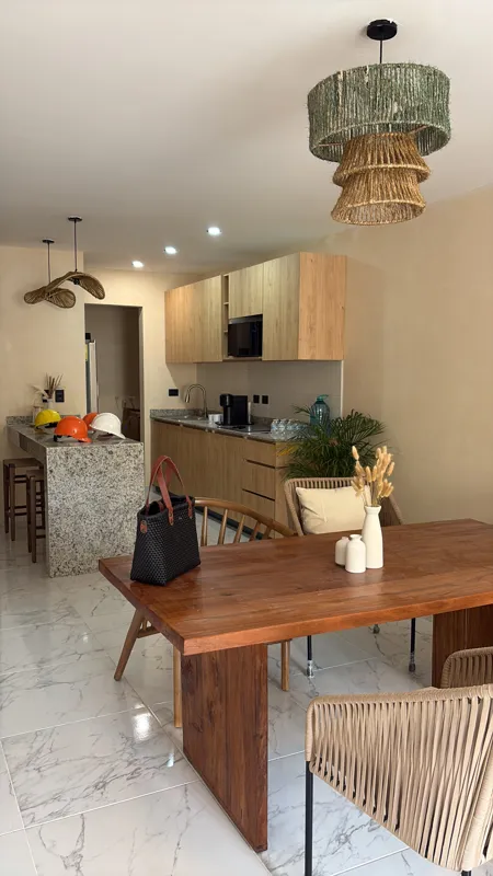 A wide shot of a modern, fully furnished kitchen and dining area in a Gobernador 28 showroom unit, featuring light wood cabinetry, a granite breakfast bar with stools, a wooden dining table with chairs, decorative woven pendant lights, and white m...