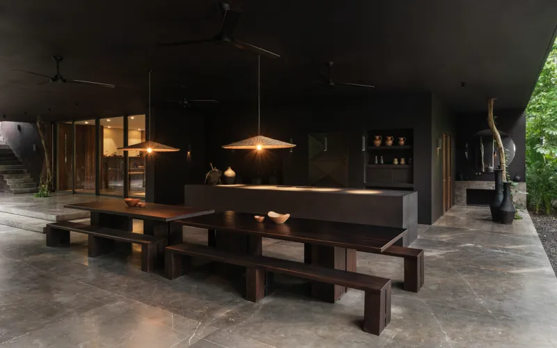 A beautifully designed outdoor lounge area at La Reserva Tulum, featuring dark walls, a tree growing through the floor, stylish dark furniture, a round coffee table, and woven pendant lights, all set against a backdrop of lush tropical foliage wit...